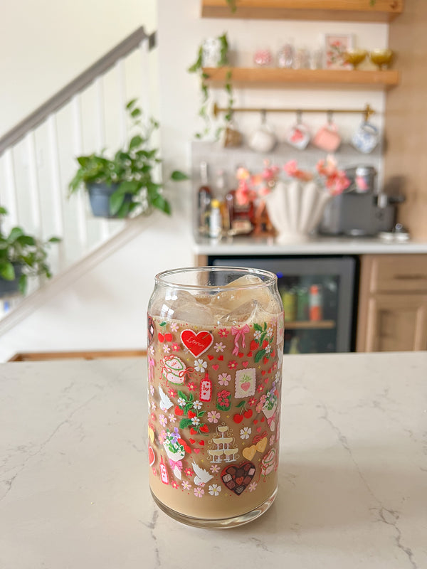 A cute 16oz glass coffee cup that is shaped like a beer can and has valentine designs around the cup in red, pink, yellow, and white. There is a heart shaped box of chocolates, champagne tower, bouquet of flowers, cherries, lockets, heart shaped cake, strawberries, bows, and flowers. The perfect galentine cup. 