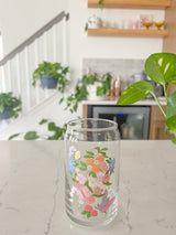 A cute 16oz glass coffee cup that is shaped like a beer can and has a butterfly garden design on the front with oranges, butterflies, pink and white flowers, and purple and blue flowers. 