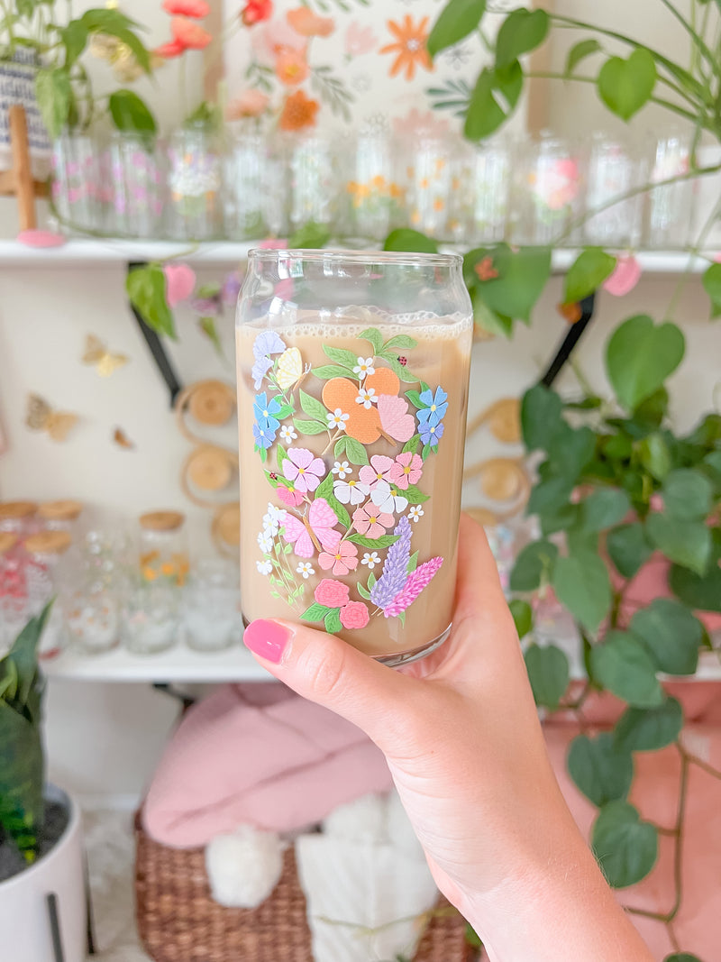 A cute 16oz glass coffee cup that is shaped like a beer can and has a butterfly garden design on the front with oranges, butterflies, pink and white flowers, and purple and blue flowers. 