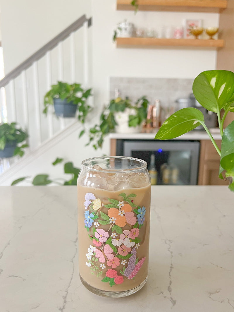 A cute 16oz glass coffee cup that is shaped like a beer can and has a butterfly garden design on the front with oranges, butterflies, pink and white flowers, and purple and blue flowers. 