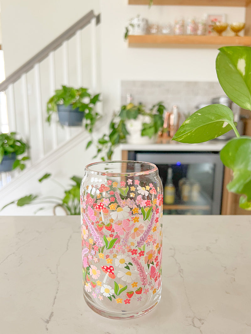 A cute 16oz glass coffee cup that is shaped like a beer can and has long purple flowers, white daisies, mushrooms, pink butterflies, and small flowers in a pattern wrapped around the glass. 