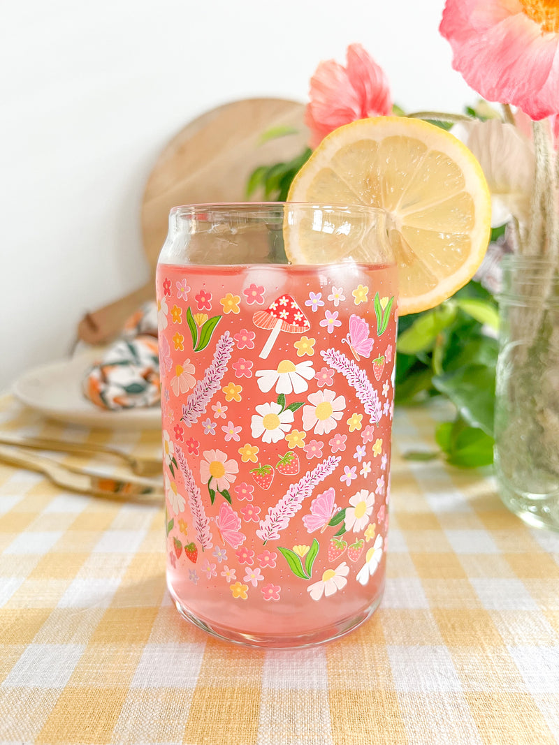 A cute 16oz glass coffee cup that is shaped like a beer can and has long purple flowers, white daisies, mushrooms, pink butterflies, and small flowers in a pattern wrapped around the glass. 