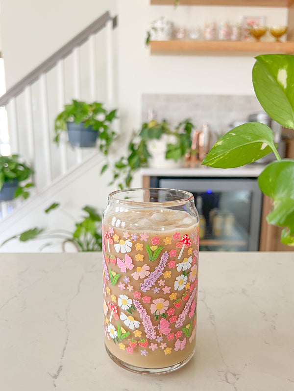 A cute 16oz glass coffee cup that is shaped like a beer can and has long purple flowers, white daisies, mushrooms, pink butterflies, and small flowers in a pattern wrapped around the glass. 