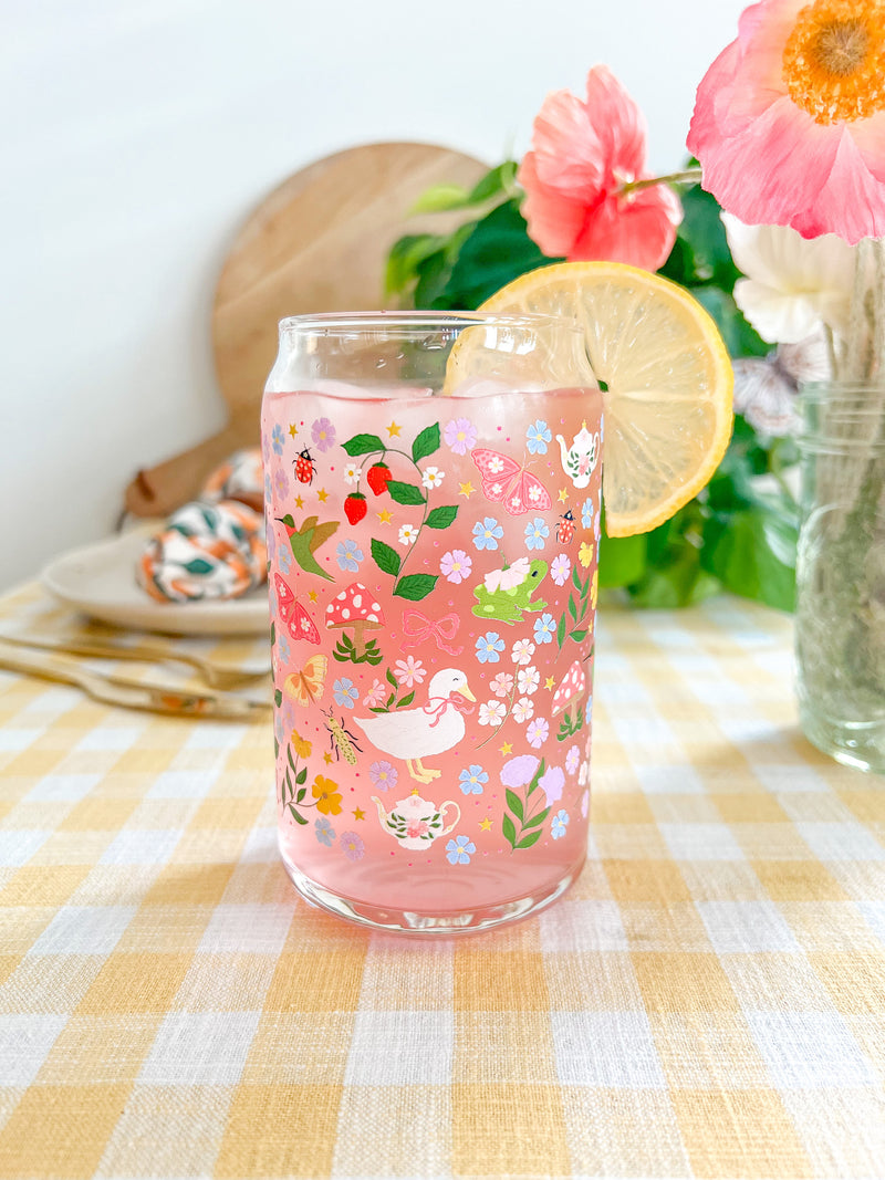 A cute 16oz glass coffee cup that is shaped like a beer can and has florals, butterflies, frogs, tea pot, bows, hummingbird, and duck with some mushrooms, strawberries, and beetles all in a repeating pattern around the glass. Color theme is purple, pink, blue and green. 
