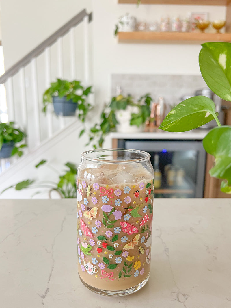 A cute 16oz glass coffee cup that is shaped like a beer can and has florals, butterflies, frogs, tea pot, bows, hummingbird, and duck with some mushrooms, strawberries, and beetles all in a repeating pattern around the glass. Color theme is purple, pink, blue and green. 