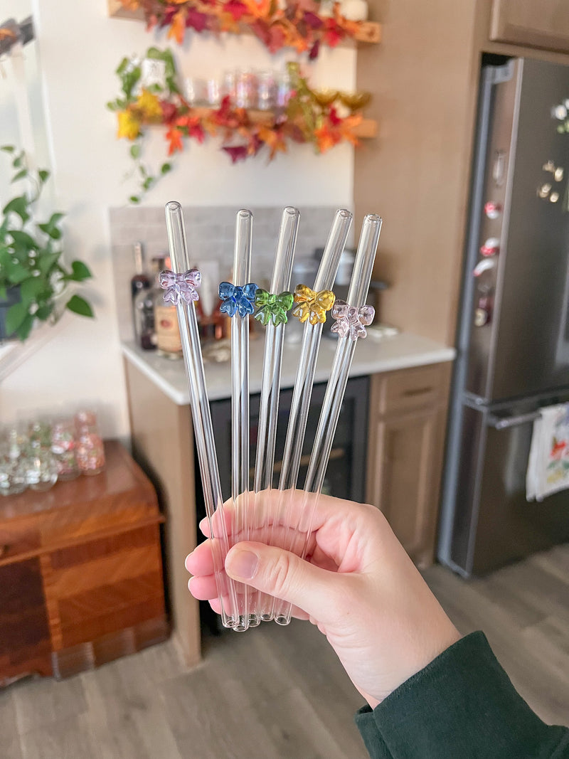 These are five different glass straws that have a bent top and bow charms on each one in pink, blue, green, yellow, and purple