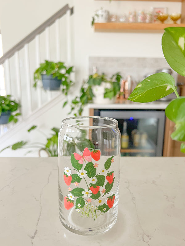 A cute 16oz glass coffee cup that is shaped like a beer can and has a design on the front that is strawberries with vines, leaves, white daises, small pink flowers and a pink butterfly. 