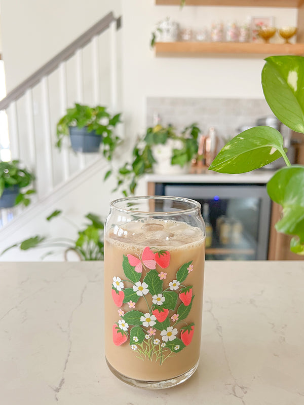 A cute 16oz glass coffee cup that is shaped like a beer can and has a design on the front that is strawberries with vines, leaves, white daises, small pink flowers and a pink butterfly. 