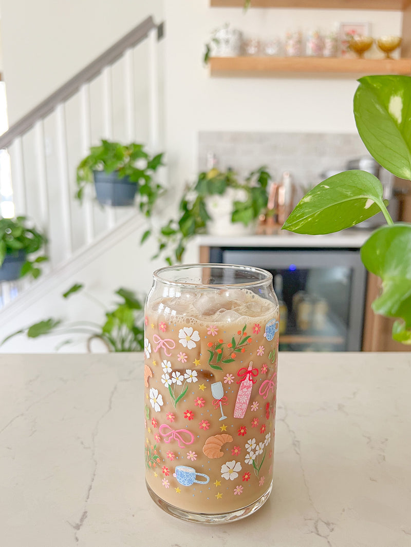 A cute 16oz glass coffee cup that is shaped like a beer can and has florals, bows, wine glass and rose bottle, tea cup, croissant, oranges, tomatoes, cake, espresso martini, pears and stars all around the glass in a repeating pattern. Color theme is pink, white, and green with a hint of blue and yellow. 