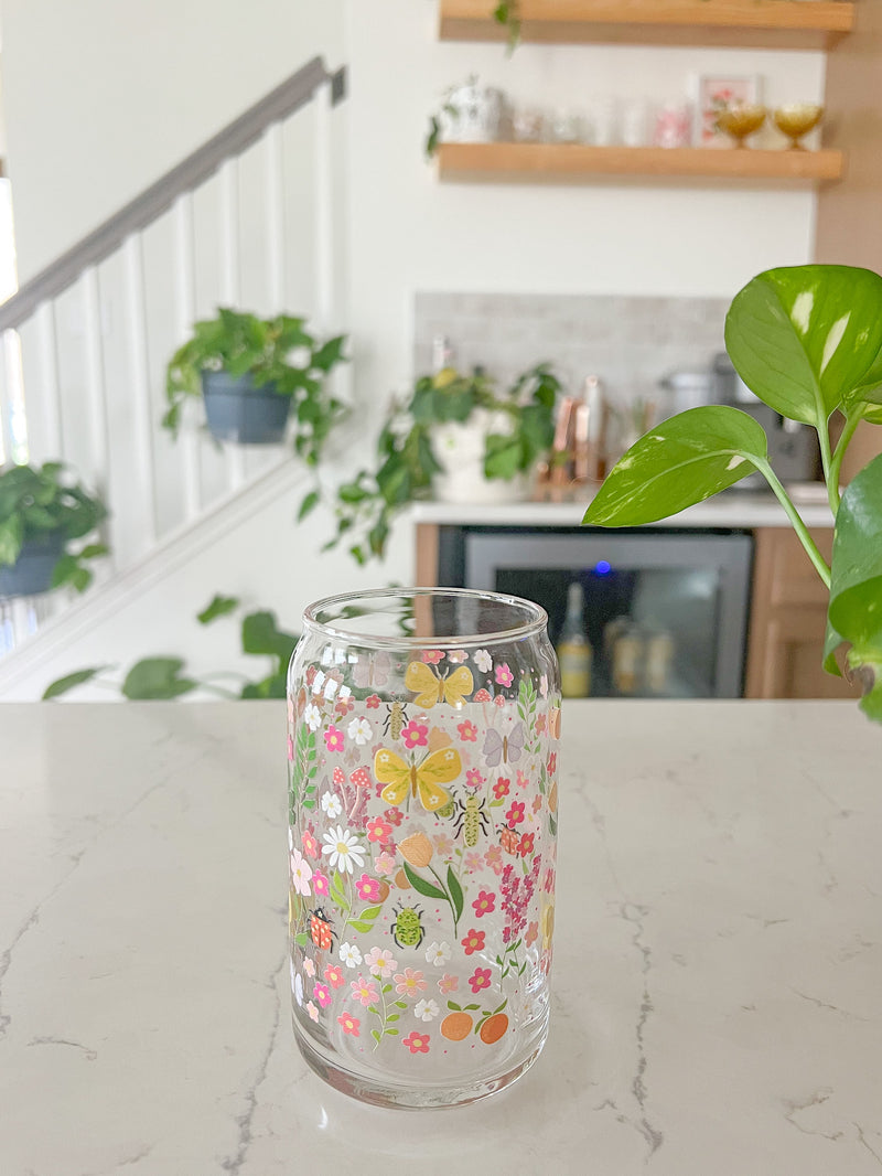 A cute 16oz glass coffee cup that is shaped like a beer can and has florals, beetles, butterflies, oranges, tulips, lilacs, ladybugs, and mushrooms in a repeating pattern around the glass. The color theme is pink, orange, and green. 