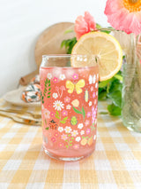 A cute 16oz glass coffee cup that is shaped like a beer can and has florals, beetles, butterflies, oranges, tulips, lilacs, ladybugs, and mushrooms in a repeating pattern around the glass. The color theme is pink, orange, and green. 