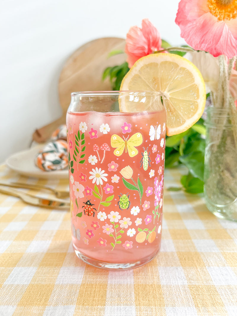 A cute 16oz glass coffee cup that is shaped like a beer can and has florals, beetles, butterflies, oranges, tulips, lilacs, ladybugs, and mushrooms in a repeating pattern around the glass. The color theme is pink, orange, and green. 