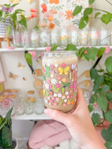 A cute 16oz glass coffee cup that is shaped like a beer can and has florals, beetles, butterflies, oranges, tulips, lilacs, ladybugs, and mushrooms in a repeating pattern around the glass. The color theme is pink, orange, and green. 