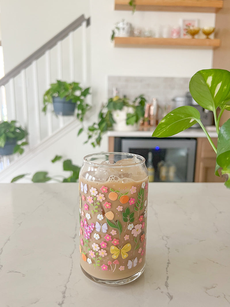 A cute 16oz glass coffee cup that is shaped like a beer can and has florals, beetles, butterflies, oranges, tulips, lilacs, ladybugs, and mushrooms in a repeating pattern around the glass. The color theme is pink, orange, and green. 