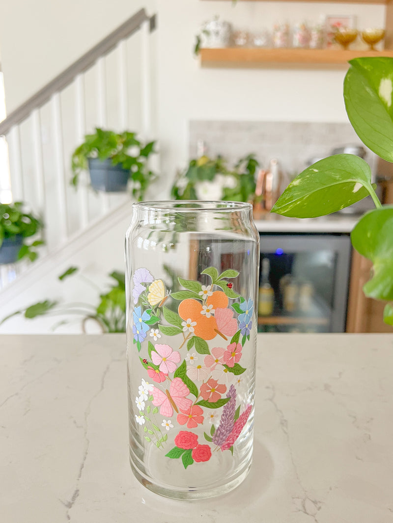 A cute 20oz glass coffee cup that is shaped like a beer can and has a butterfly garden design on the front with oranges, butterflies, pink and white flowers, and purple and blue flowers. 