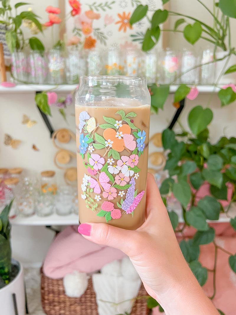 A cute 20oz glass coffee cup that is shaped like a beer can and has a butterfly garden design on the front with oranges, butterflies, pink and white flowers, and purple and blue flowers. 