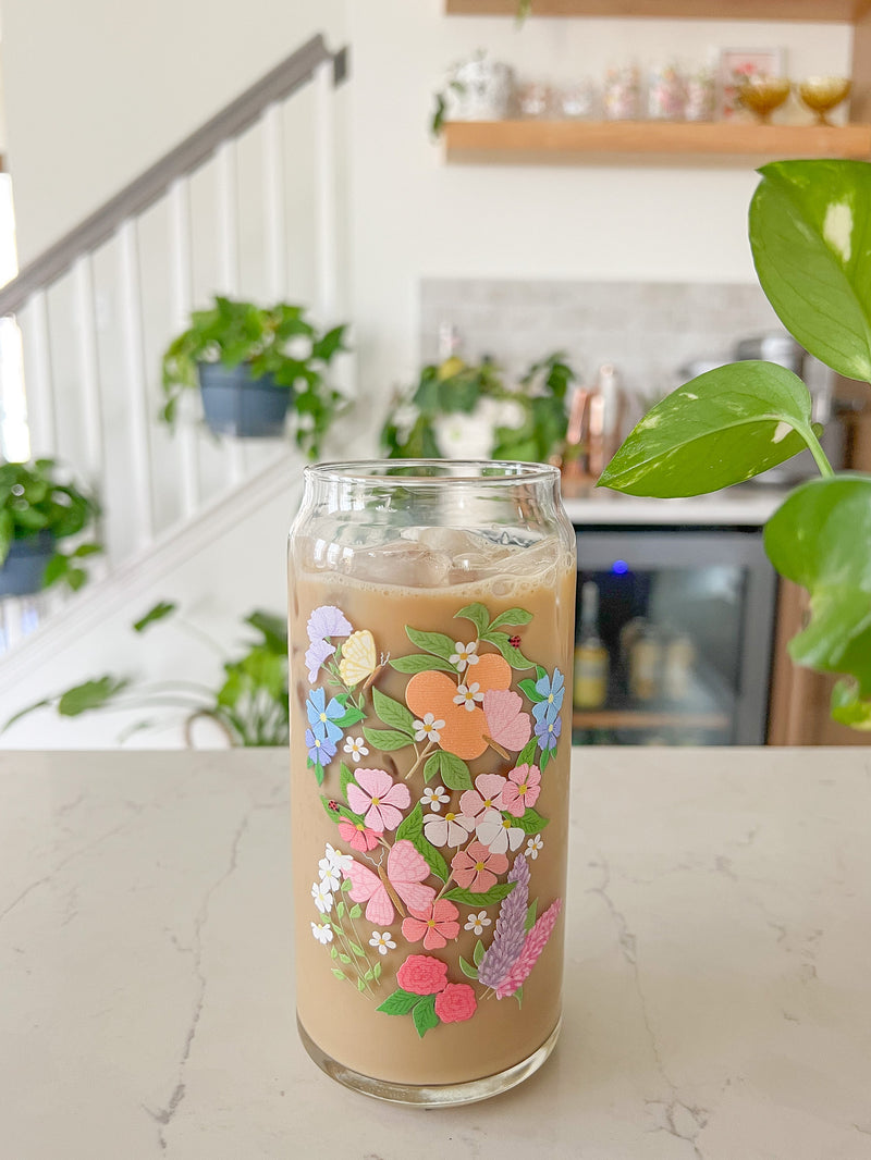 A cute 20oz glass coffee cup that is shaped like a beer can and has a butterfly garden design on the front with oranges, butterflies, pink and white flowers, and purple and blue flowers. 