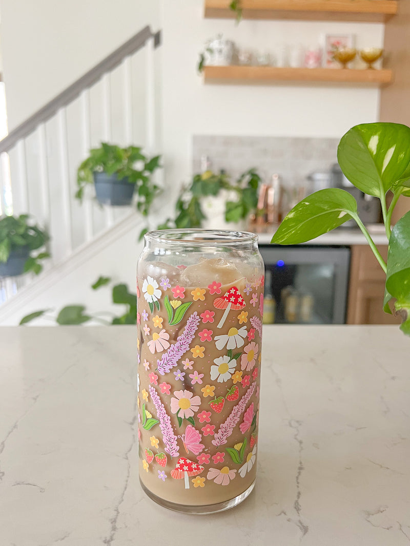 A cute 20oz glass coffee cup that is shaped like a beer can and has long purple flowers, white daisies, mushrooms, pink butterflies, and small flowers in a pattern wrapped around the glass. 