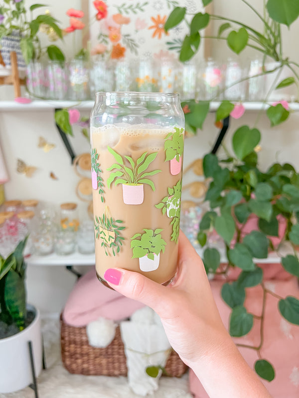 A cute 20oz glass coffee cup that is shaped like a beer can and has different house plants around the glass in green, with pink and orange pots. Some are hanging plants and some are potted plants. 