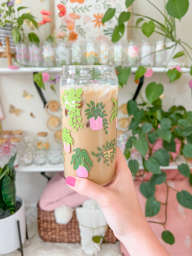 A cute 20oz glass coffee cup that is shaped like a beer can and has different house plants around the glass in green, with pink and orange pots. Some are hanging plants and some are potted plants. 