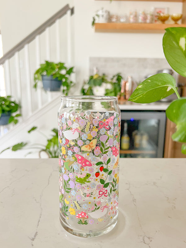 A cute 20oz glass coffee cup that is shaped like a beer can and has florals, butterflies, frogs, tea pot, bows, hummingbird, and duck with some mushrooms, strawberries, and beetles all in a repeating pattern around the glass. Color theme is purple, pink, blue and green. 