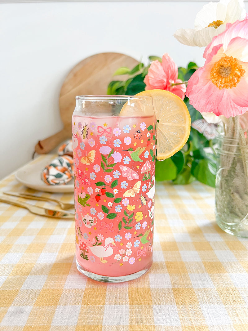 A cute 20oz glass coffee cup that is shaped like a beer can and has florals, butterflies, frogs, tea pot, bows, hummingbird, and duck with some mushrooms, strawberries, and beetles all in a repeating pattern around the glass. Color theme is purple, pink, blue and green. 