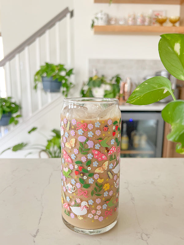 A cute 20oz glass coffee cup that is shaped like a beer can and has florals, butterflies, frogs, tea pot, bows, hummingbird, and duck with some mushrooms, strawberries, and beetles all in a repeating pattern around the glass. Color theme is purple, pink, blue and green. 