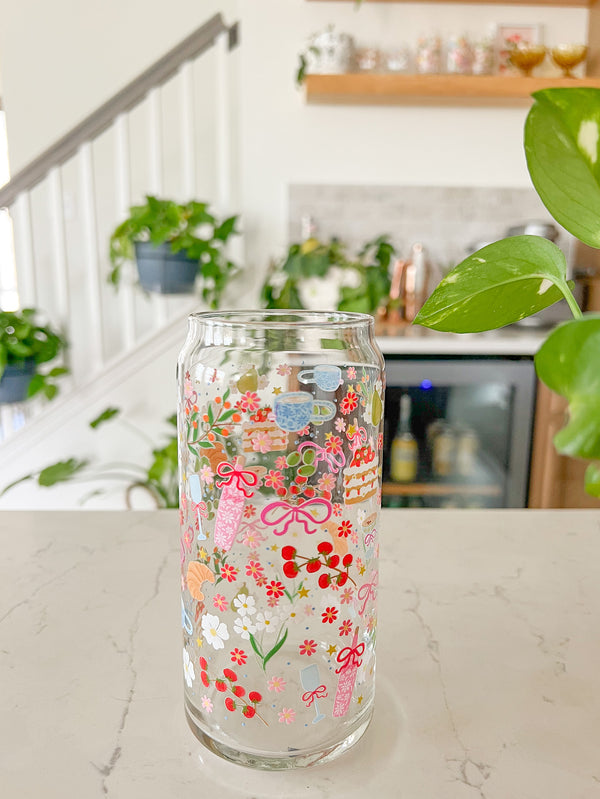 A cute 20oz glass coffee cup that is shaped like a beer can and has florals, bows, wine glass and rose bottle, tea cup, croissant, oranges, tomatoes, cake, espresso martini, pears and stars all around the glass in a repeating pattern. Color theme is pink, white, and green with a hint of blue and yellow. 