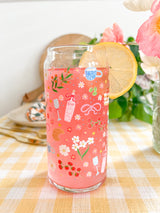 A cute 20oz glass coffee cup that is shaped like a beer can and has florals, bows, wine glass and rose bottle, tea cup, croissant, oranges, tomatoes, cake, espresso martini, pears and stars all around the glass in a repeating pattern. Color theme is pink, white, and green with a hint of blue and yellow. 