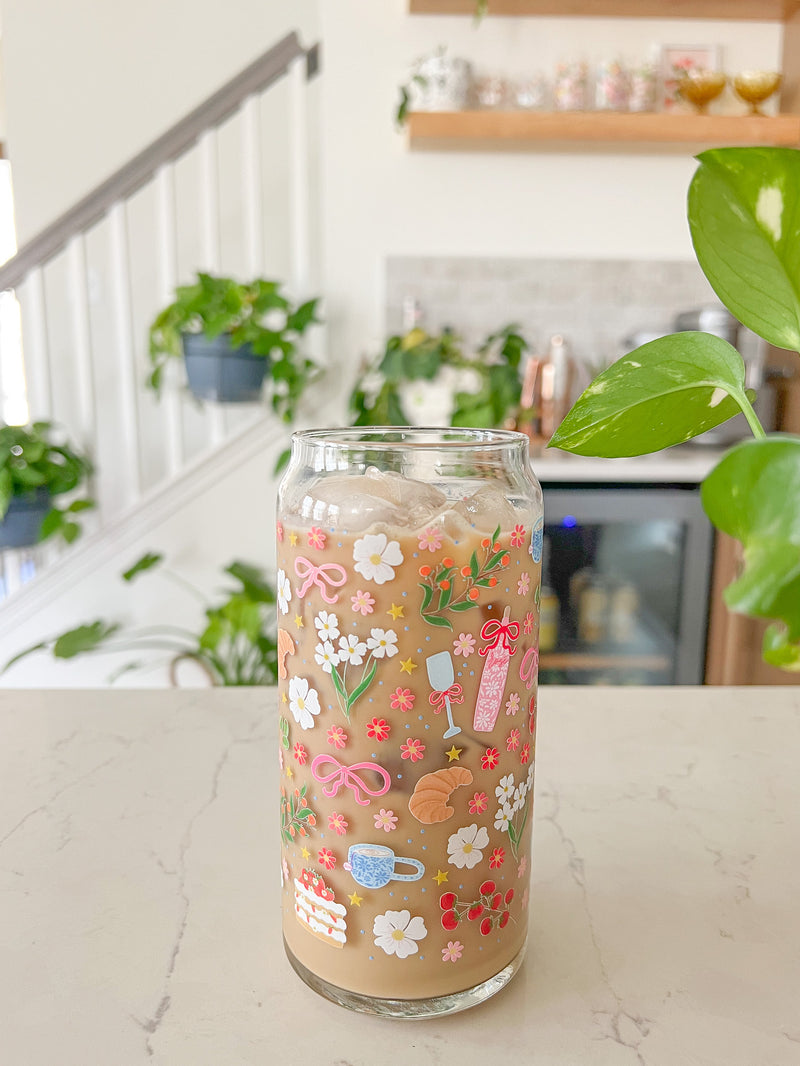 A cute 20oz glass coffee cup that is shaped like a beer can and has florals, bows, wine glass and rose bottle, tea cup, croissant, oranges, tomatoes, cake, espresso martini, pears and stars all around the glass in a repeating pattern. Color theme is pink, white, and green with a hint of blue and yellow. 