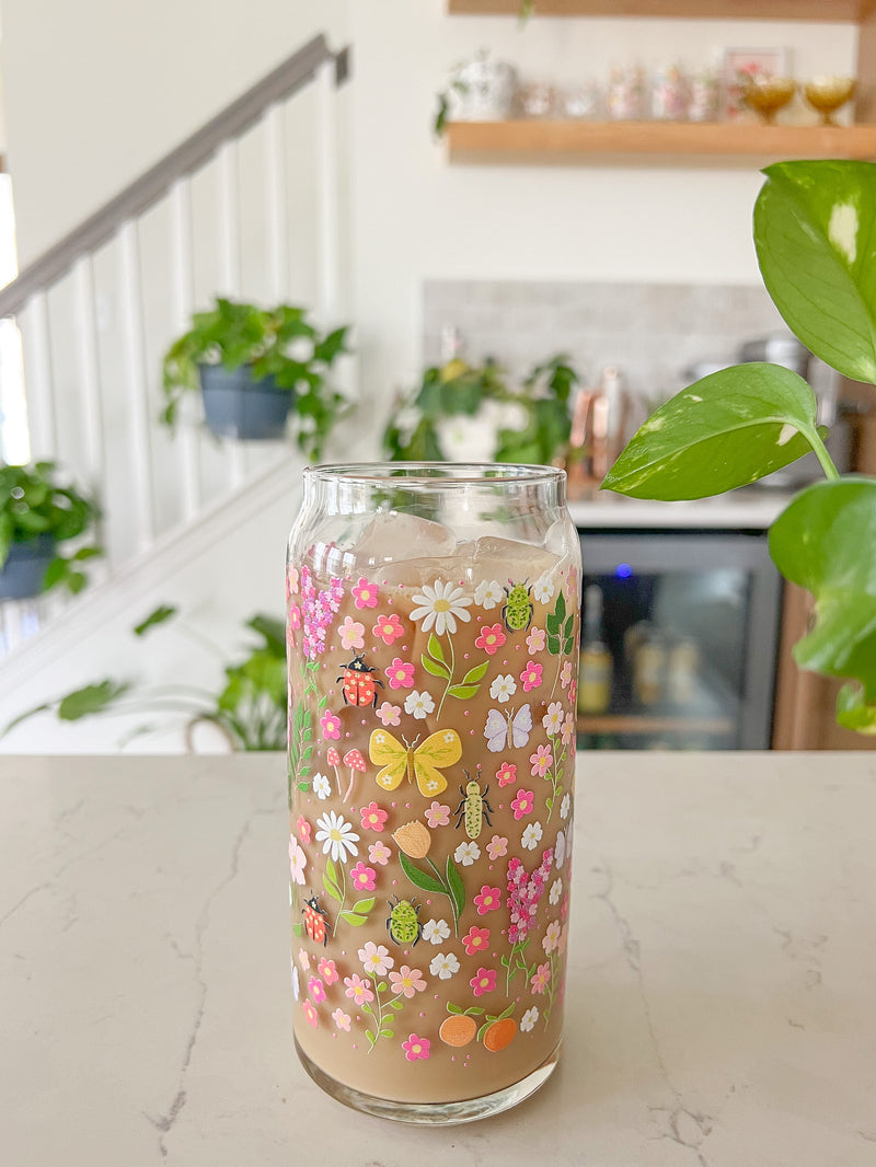 A cute 20oz glass coffee cup that is shaped like a beer can and has florals, beetles, butterflies, oranges, tulips, lilacs, ladybugs, and mushrooms in a repeating pattern around the glass. The color theme is pink, orange, and green. 