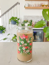 A cute 20oz libbey beer can glass coffee cup that is shaped like a beer can and has a design on the front that is strawberries with vines, leaves, white daises, small pink flowers and a pink butterfly. 