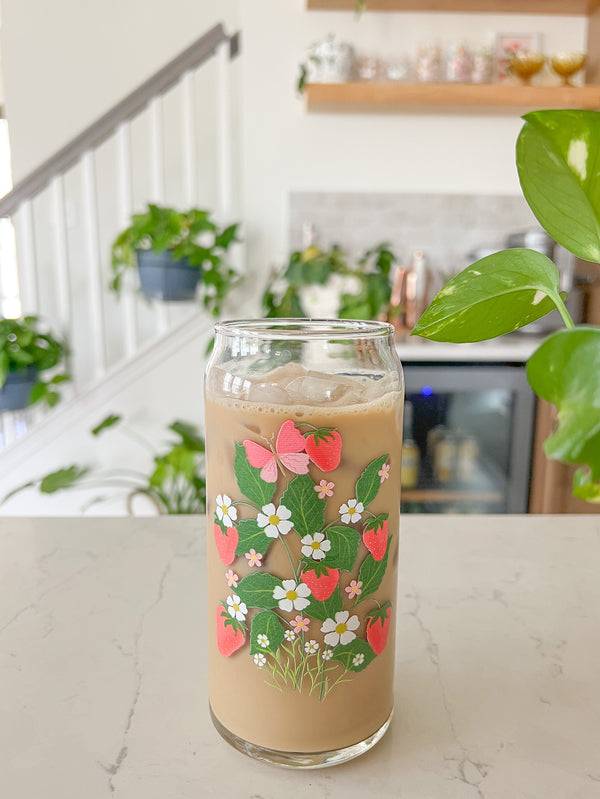 A cute 20oz libbey beer can glass coffee cup that is shaped like a beer can and has a design on the front that is strawberries with vines, leaves, white daises, small pink flowers and a pink butterfly. 