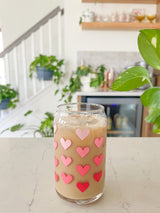 A cute 16oz glass coffee cup that is shaped like a beer can and has different shade of pink hearts around the glass. 