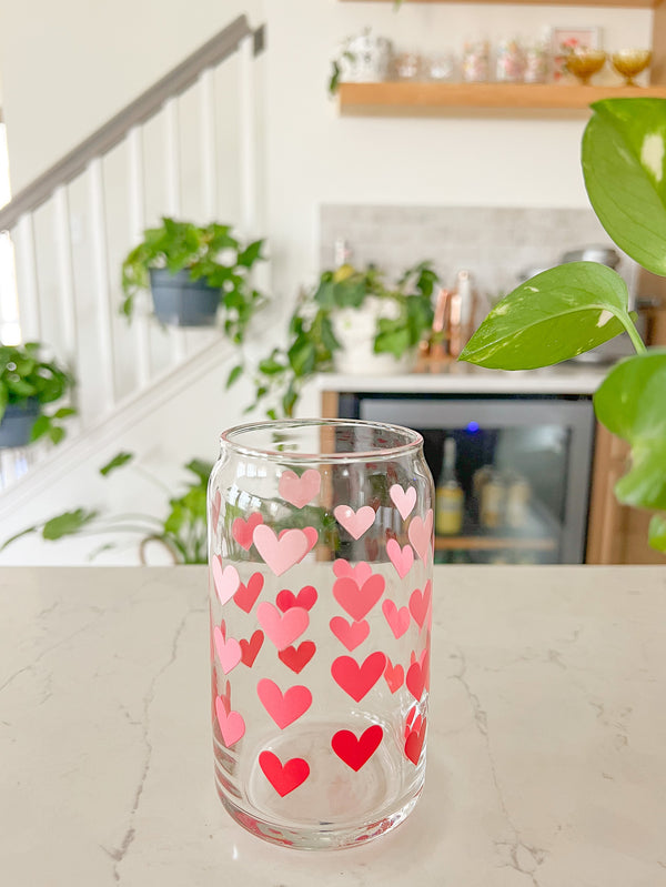 A cute 16oz glass coffee cup that is shaped like a beer can and has different shade of pink hearts around the glass. 