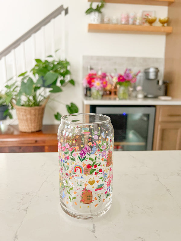 A cute 16oz glass coffee cup that is shaped like a beer can and has florals, locket and key, butterflies, secret garden door, mushrooms, books, stars, rainbows, bird bath, and tulips in a repeating pattern around the glass. Colors are pink, purple, blue and green. 