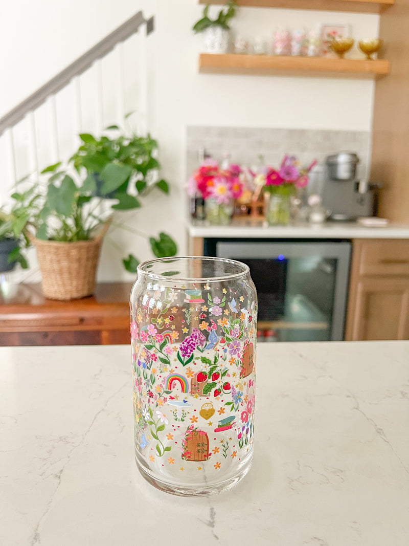 A cute 16oz glass coffee cup that is shaped like a beer can and has florals, locket and key, butterflies, secret garden door, mushrooms, books, stars, rainbows, bird bath, and tulips in a repeating pattern around the glass. Colors are pink, purple, blue and green. 