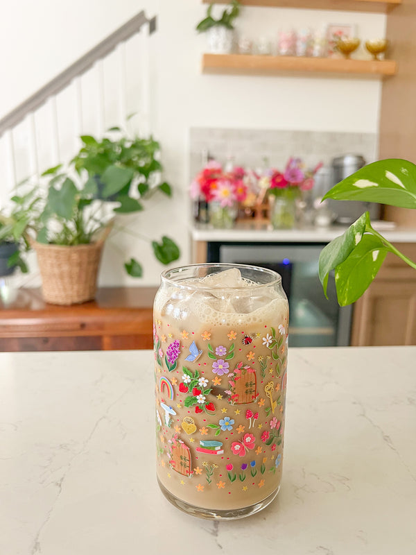 A cute 16oz glass coffee cup that is shaped like a beer can and has florals, locket and key, butterflies, secret garden door, mushrooms, books, stars, rainbows, bird bath, and tulips in a repeating pattern around the glass. Colors are pink, purple, blue and green. 