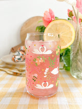 A cute 16oz glass coffee cup that is shaped like a beer can and has swans in water, lemons, oranges on branches, pink bows, and florals in a repeating pattern around the glass. 