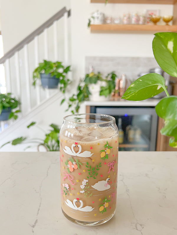 A cute 16oz glass coffee cup that is shaped like a beer can and has swans in water, lemons, oranges on branches, pink bows, and florals in a repeating pattern around the glass. 