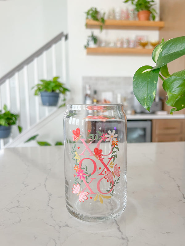 A cute 16oz glass coffee cup that is shaped like a beer can and has a design on the front of the glass that say "XO OX XO" and has florals and butterflies with heart wings around the letters. The colors are hot pink, light pink and pops of yellow and white. 