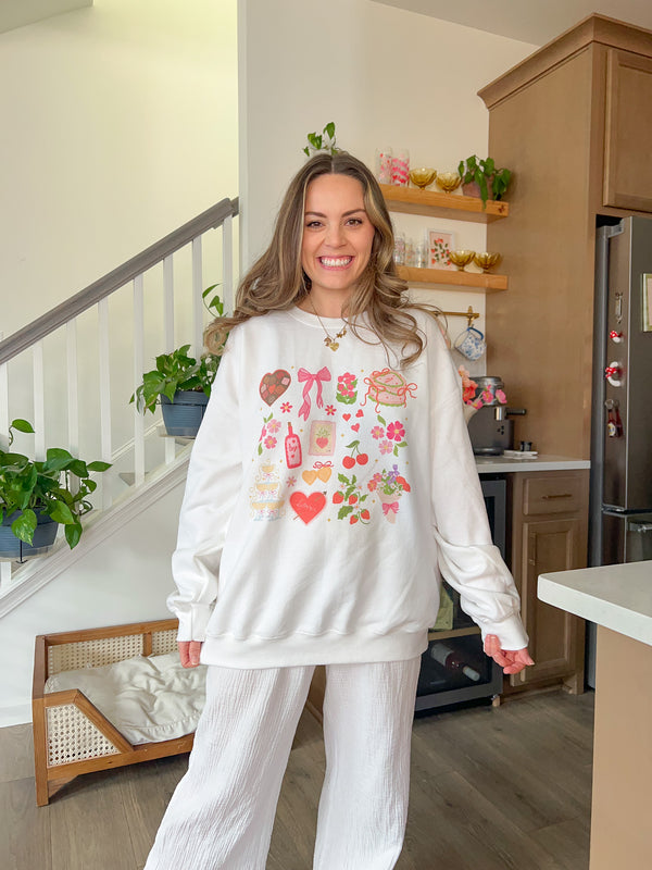 This is a cute sweatshirt that has valentine party essentials on the front. Heart shaped box of chocolates, bows, flowers, wine bottle, heart shaped cake, flower bouquet, champagne tower, lockets and more. Perfect for a a galentine party.