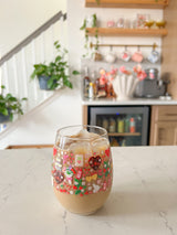 A cute wine glass cup that has valentine designs around the cup in red, pink, yellow, and white. There is a heart shaped box of chocolates, champagne tower, bouquet of flowers, cherries, lockets, heart shaped cake, strawberries, bows, and flowers. The perfect galentine cup. 