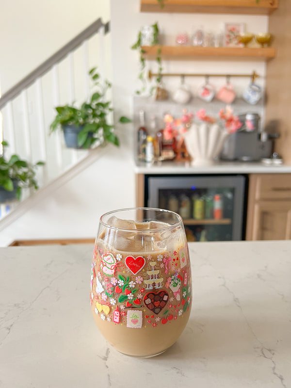 A cute wine glass cup that has valentine designs around the cup in red, pink, yellow, and white. There is a heart shaped box of chocolates, champagne tower, bouquet of flowers, cherries, lockets, heart shaped cake, strawberries, bows, and flowers. The perfect galentine cup. 