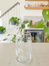 A cute 16oz glass coffee cup that is shaped like a beer can and has different types of house plants around the entire glass. Each houseplant is white and an outline. They are all in different unique pots. 