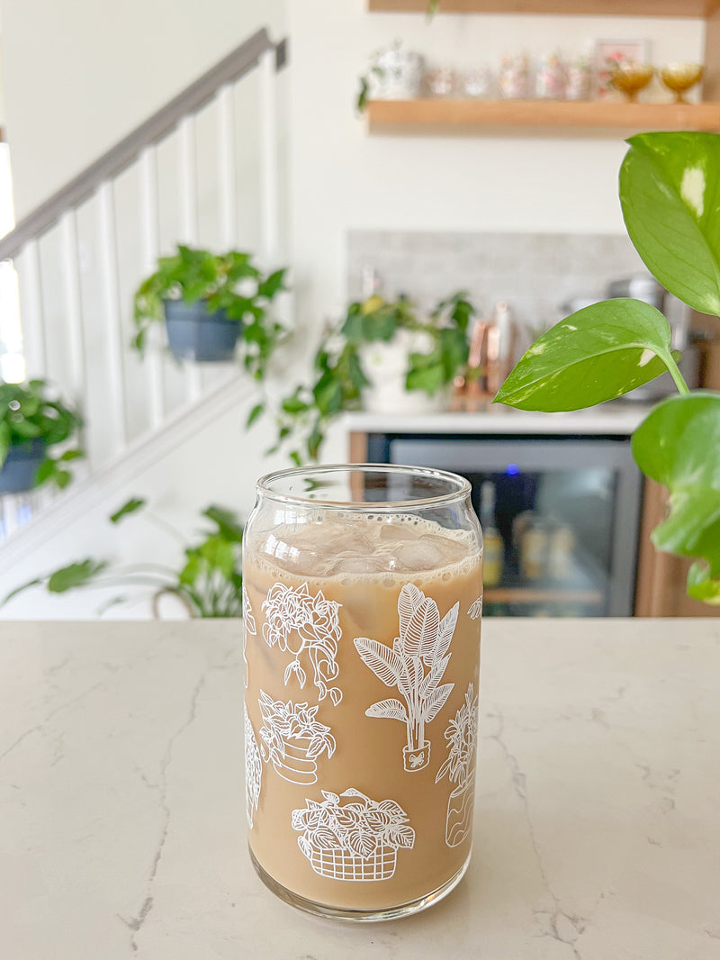 A cute 16oz glass coffee cup that is shaped like a beer can and has different types of house plants around the entire glass. Each houseplant is white and an outline. They are all in different unique pots. 