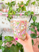 A cute 16oz glass coffee cup that is shaped like a beer can and has long purple flowers, white daisies, mushrooms, pink butterflies, and small flowers in a pattern wrapped around the glass. 