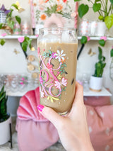 A cute 20oz libbey beer can glass coffee cup that is shaped like a beer can and has a design on the front of the glass that say "XO OX XO" and has florals and butterflies with heart wings around the letters. The colors are hot pink, light pink and pops of yellow and white. 