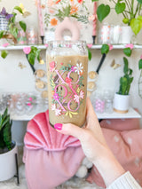 A cute 20oz libbey beer can glass coffee cup that is shaped like a beer can and has a design on the front of the glass that say "XO OX XO" and has florals and butterflies with heart wings around the letters. The colors are hot pink, light pink and pops of yellow and white. 