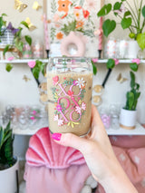 A cute 16oz glass coffee cup that is shaped like a beer can and has a design on the front of the glass that say "XO OX XO" and has florals and butterflies with heart wings around the letters. The colors are hot pink, light pink and pops of yellow and white. 